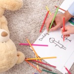 Close up of hands of father and his child are drawing picture of themselves. They are holding colored pencils. The man and boy are sitting on flooring