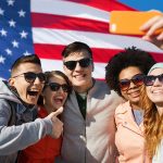 people, international friendship and technology concept - group of happy teenage friends taking selfie with smartphone and showing thumbs up over american flag background